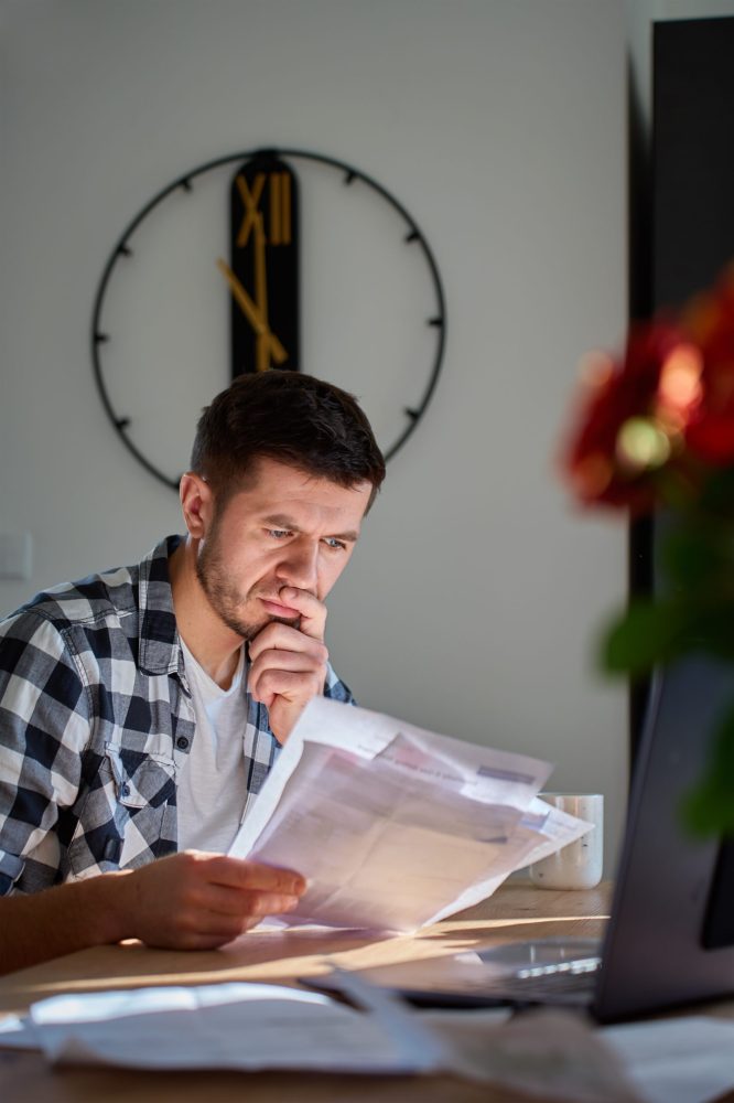 Man sitting at desk at home checking utility bills and financial papers with laptop. Concept of high household expenses, budgeting and financial stress.